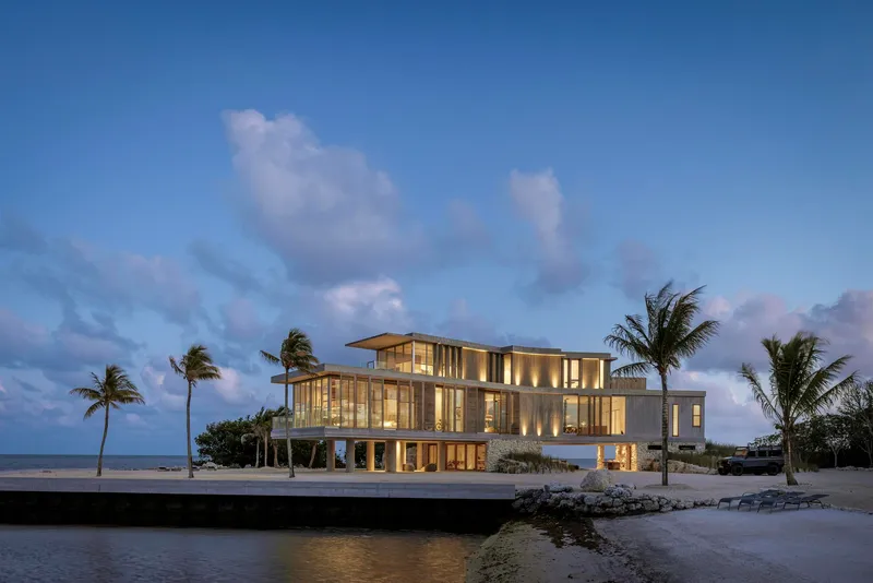 Waterside elevation showing the illuminated glass facades and concrete structure at twilight with swaying palms