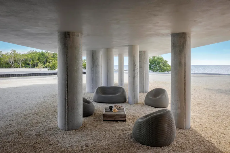 Shaded ground-floor seating area beneath cylindrical concrete columns with gravel floor and coastal view