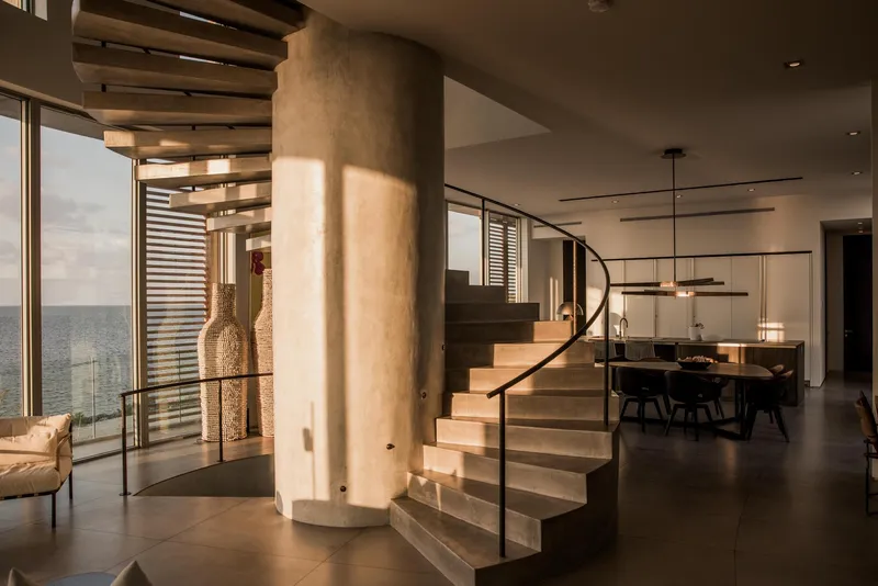 Curved concrete staircase wrapping a cylindrical column with dining area visible beyond at sunset