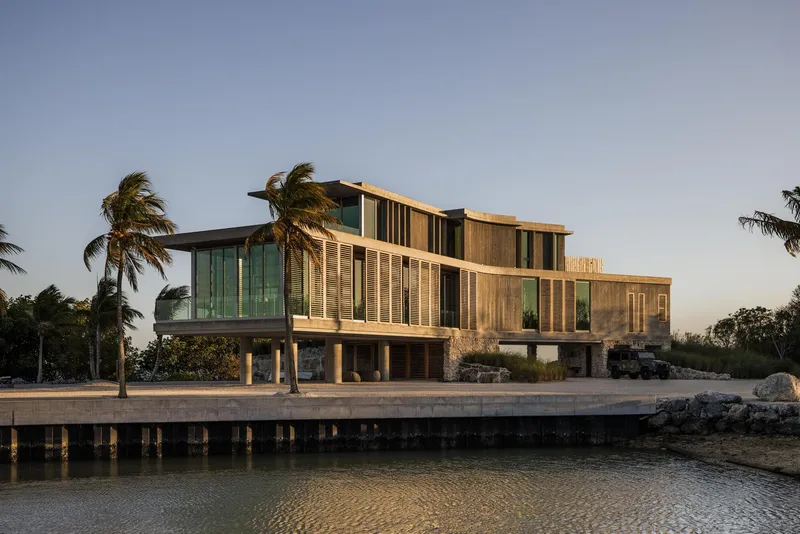 Waterfront elevation with timber and glass volumes elevated on concrete piers under swaying palm trees