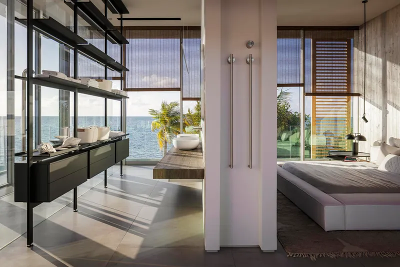 Open bedroom suite with freestanding white partition and full-height glazing framing coastal vegetation