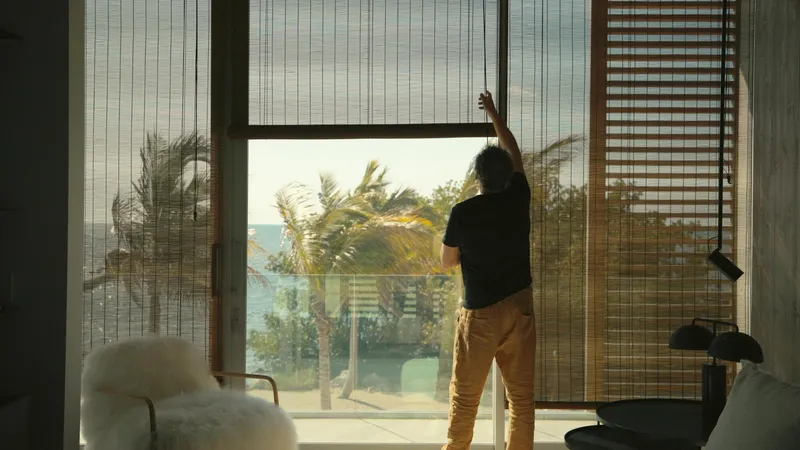 Interior view of a person adjusting bamboo blinds at floor-to-ceiling glazing overlooking palm trees and ocean
