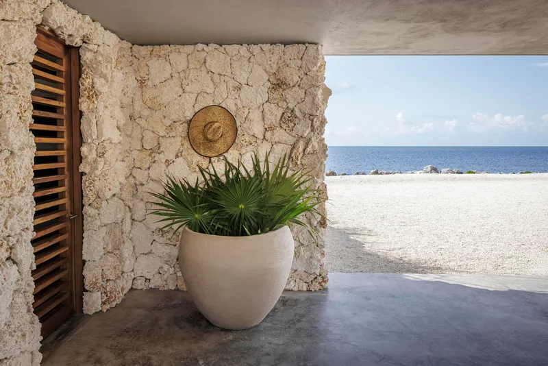 Shaded porch corner with coral stone wall, timber louver door, planted urn, and view to white gravel and ocean