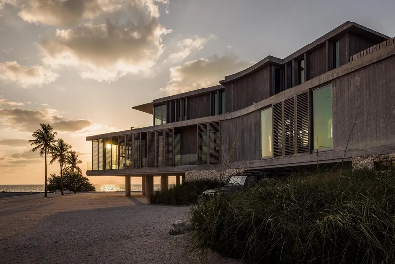 Elevated timber-clad volume on pilotis with slatted screens and cantilevered roof plane at sunset beside palm trees