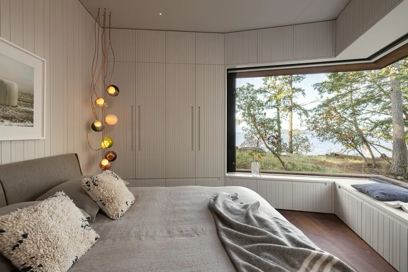 Bedroom with ribbed wall paneling and pendant lights beside a corner window framing coastal trees