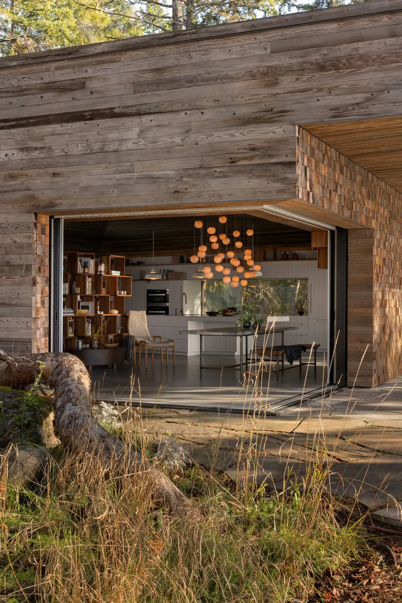 Wide opening in timber facade revealing kitchen and dining area with clustered pendant lights at dusk