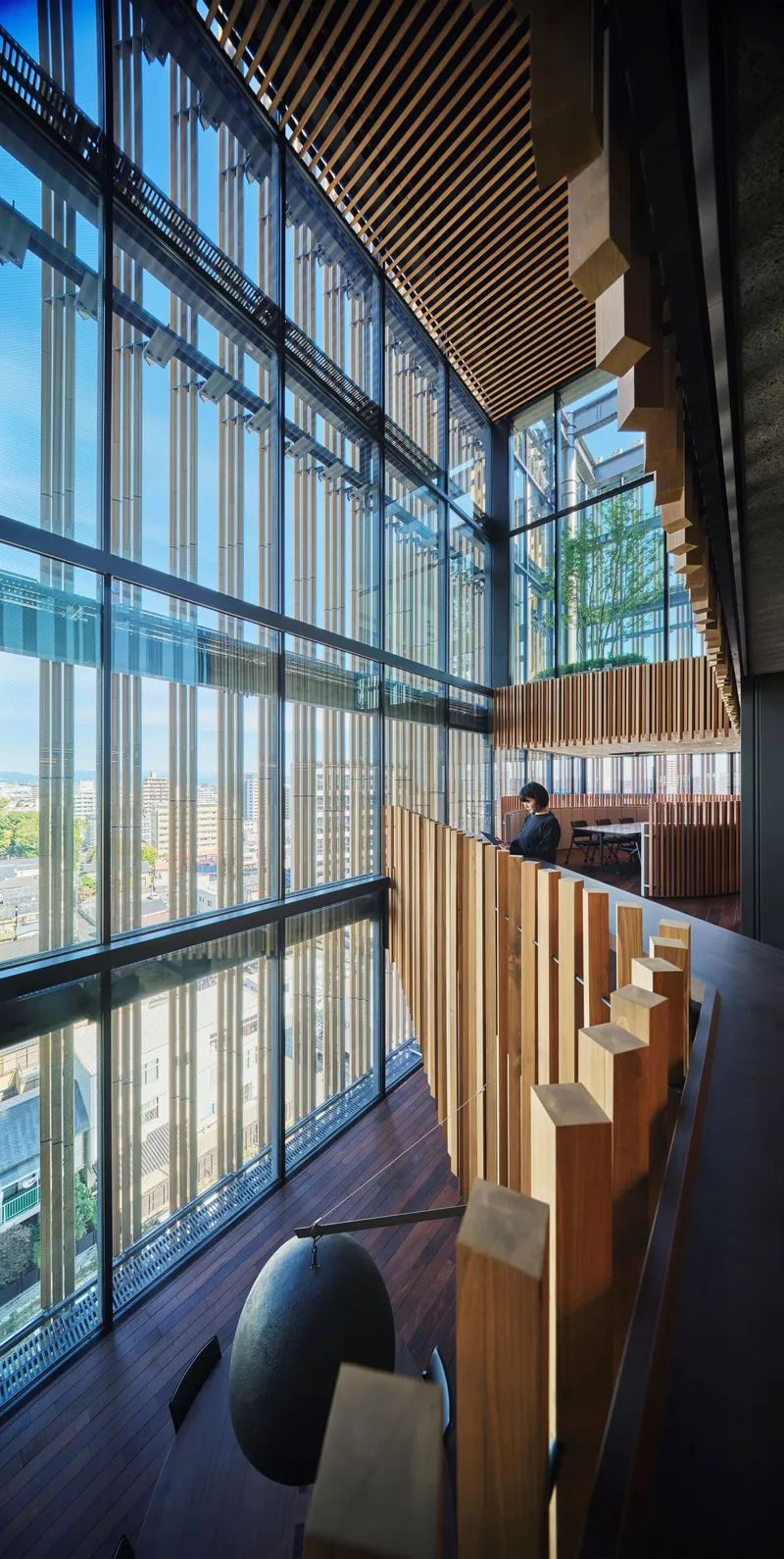 Multi-story atrium with vertical timber slat screens, glass curtain wall, and floating timber ceiling elements