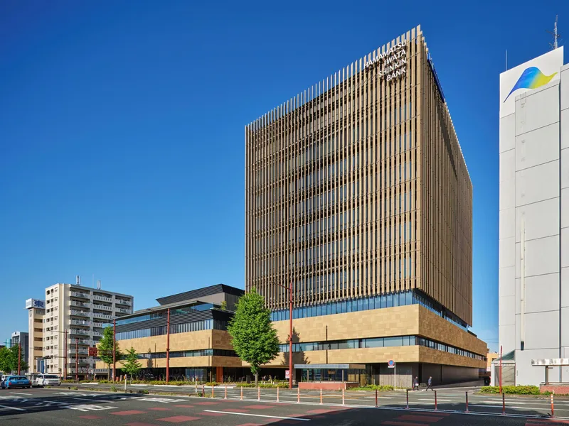Street view of the vertical timber brise-soleil facade rising above a horizontal stone base under a clear blue sky