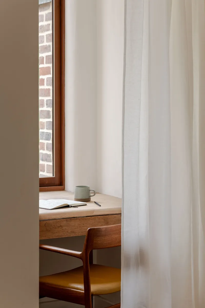 Detail of a writing desk with timber chair behind a sheer curtain beside a narrow window