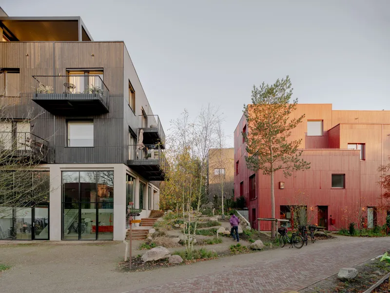 Shared courtyard with young trees and brick paving between multifamily housing volumes clad in timber and metal