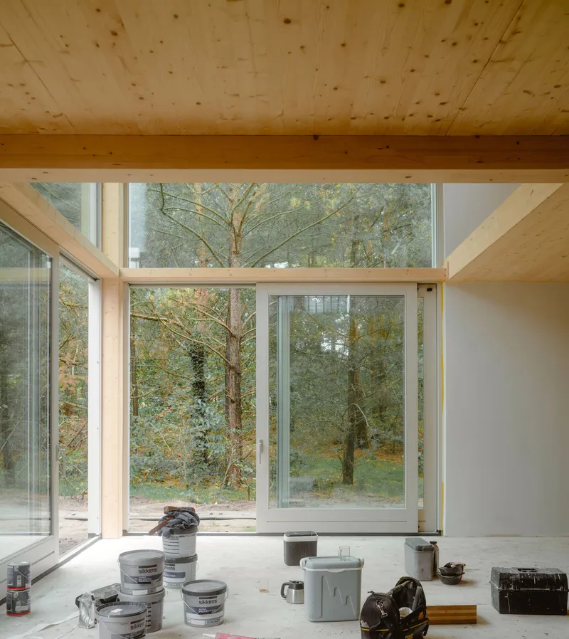 Interior view through large timber-framed windows overlooking trees in autumn with paint buckets on the floor