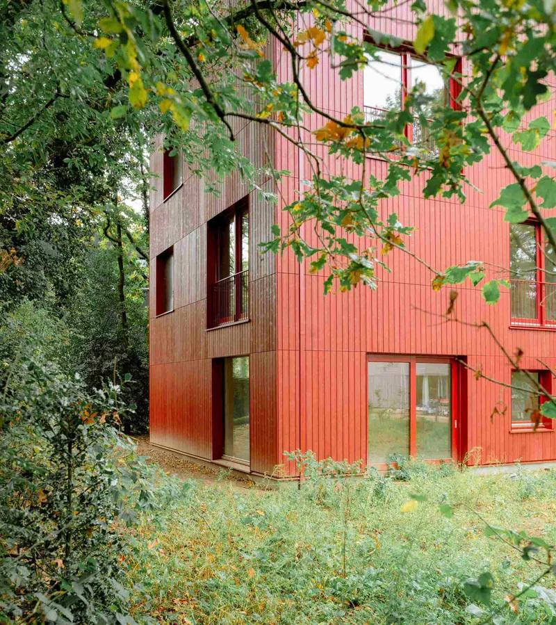Pink ribbed facade with vertical timber accent and red-framed openings framed by autumn foliage