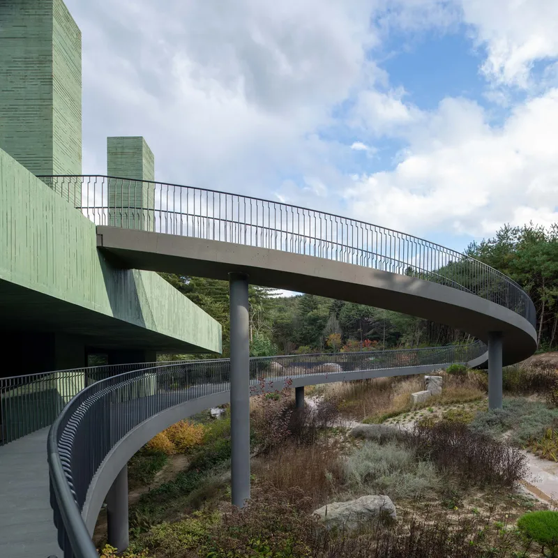 Concrete pedestrian bridge with metal railings spanning between green towers above a planted landscape