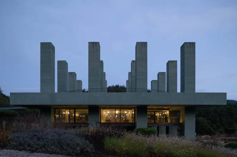 Elevated concrete slab with towers above a glazed base at dusk with interior lights glowing