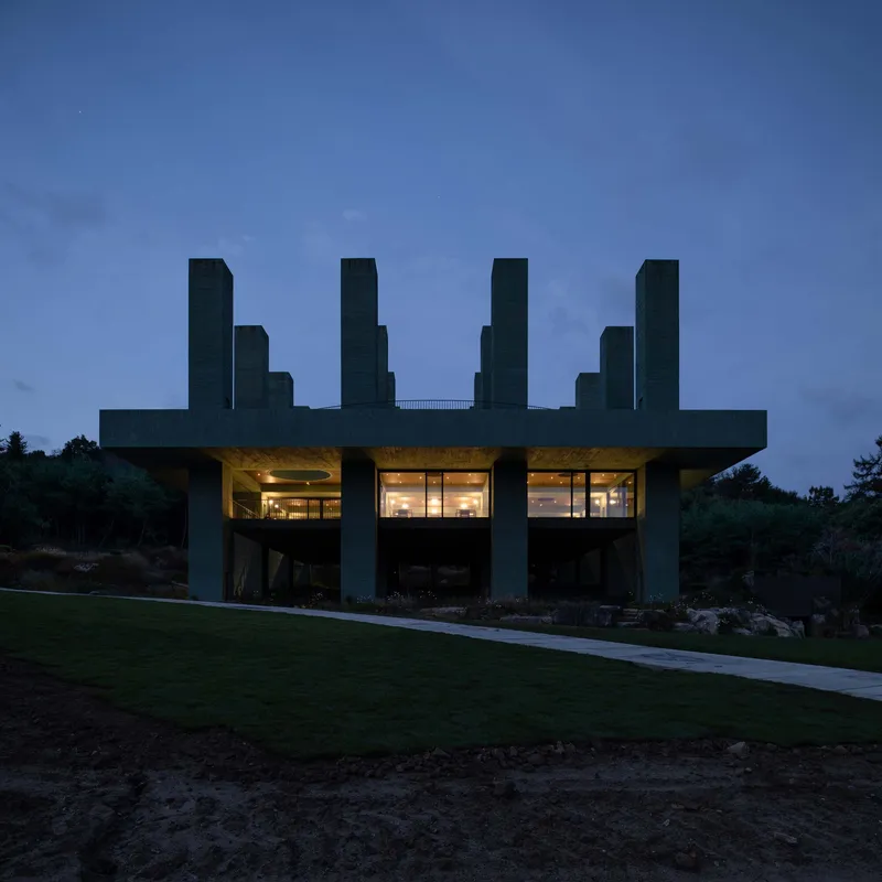 Front elevation showing illuminated interior beneath raised concrete platform with tower array at twilight