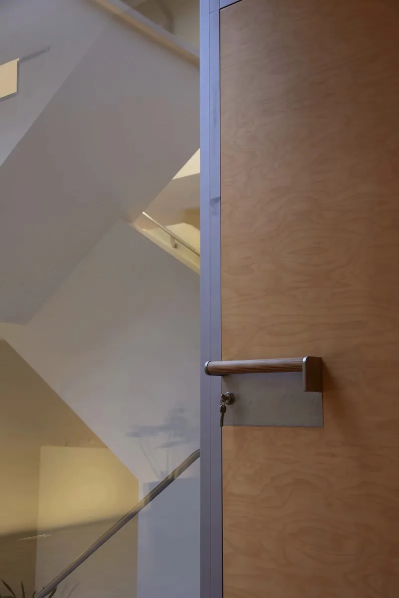Close-up of plywood door with metal handle and lock beside a white stairwell