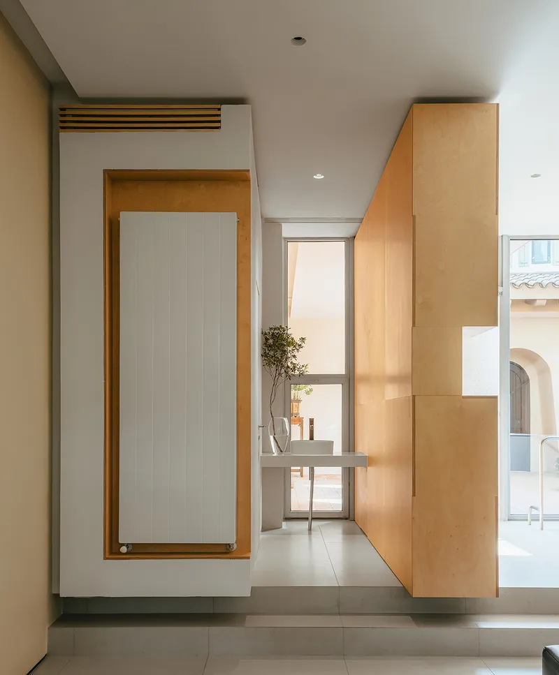 Hallway with plywood radiator panel and glass door overlooking courtyard in afternoon light