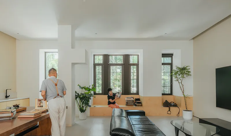 Open living space showing two people, built-in timber seating, and tall gridded windows overlooking greenery