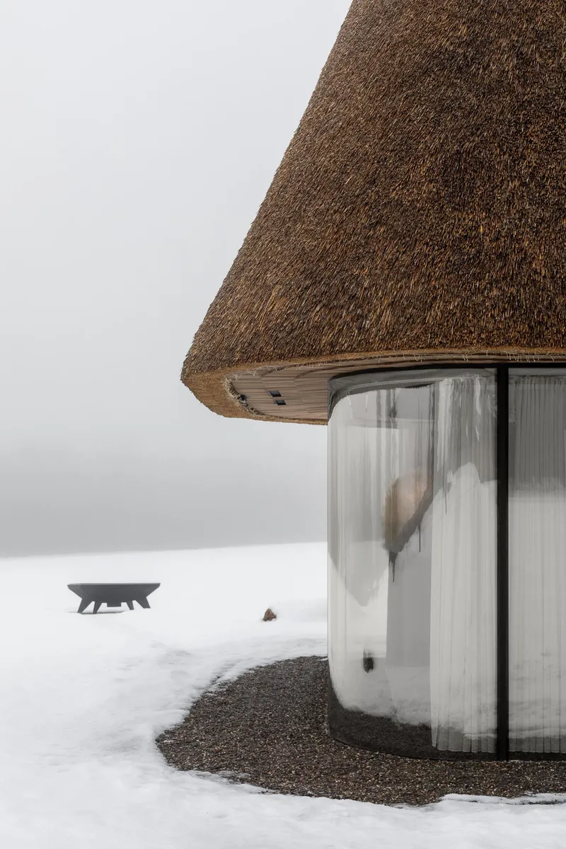 Curved glass facade beneath the thatched roof with gravel base exposed in surrounding snow