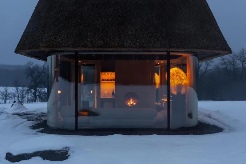 Glass-walled pavilion with conical thatched roof illuminated at dusk in a snowy landscape