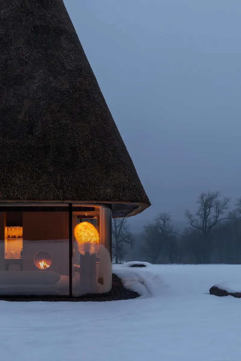 Curved glazed facade with warm interior lighting beneath a dark thatched roof at twilight