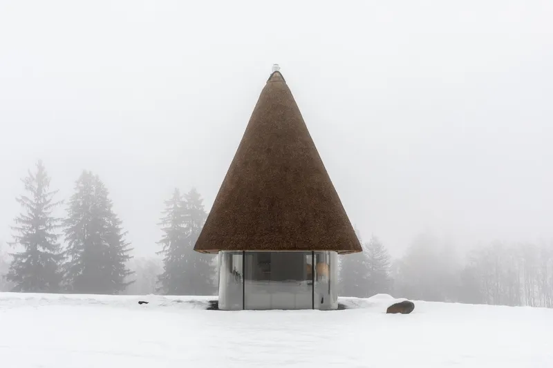 Thatched pyramidal roof pavilion standing in a snow-covered field surrounded by misty pine trees