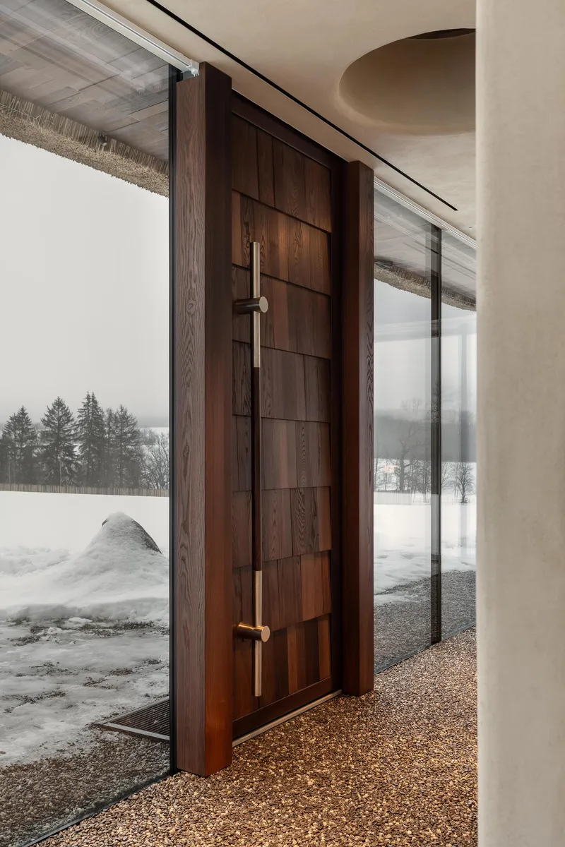 Horizontal timber-paneled entry door with bronze lever handles opening to snowy exterior