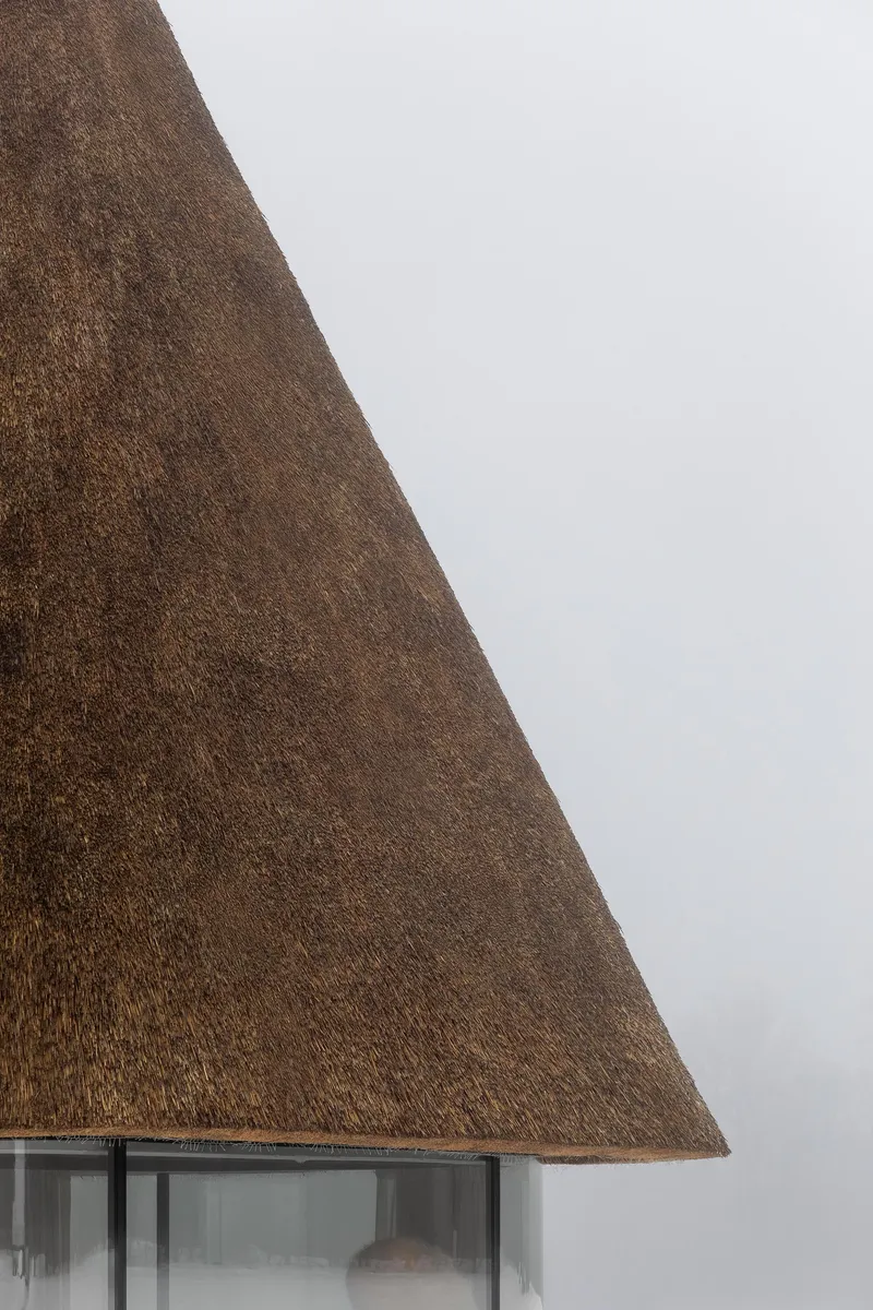 Close-up of the thatched roof edge meeting the curved glass curtain wall beneath an overcast sky