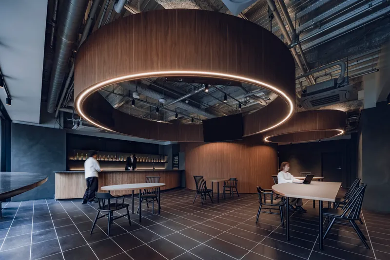 Dining area with curved wooden pods and illuminated ring fixtures as two figures move through the space