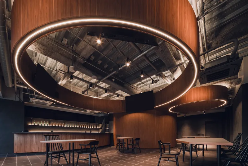 Circular wood-clad volume with integrated ring light above dining area with exposed industrial ceiling