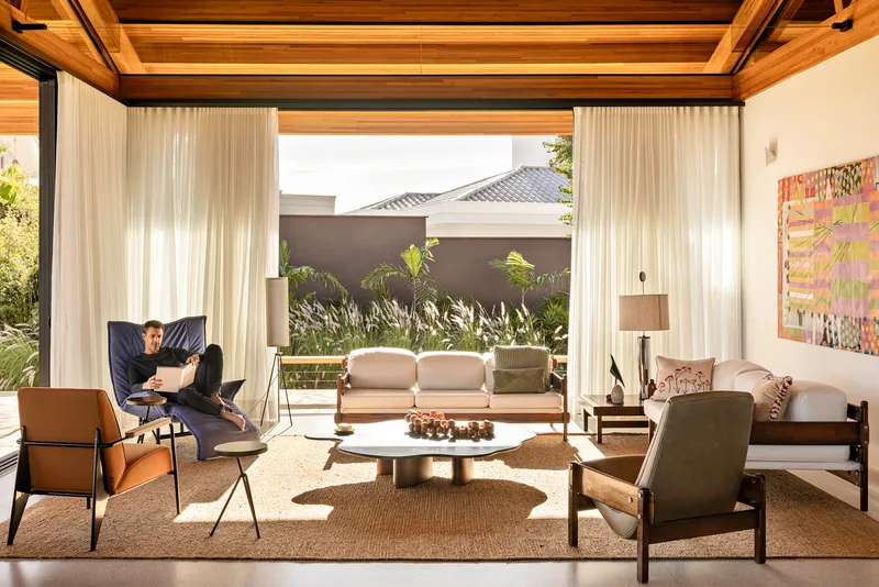 Living room framed by floor-to-ceiling curtains opening to a planted courtyard with a seated figure reading