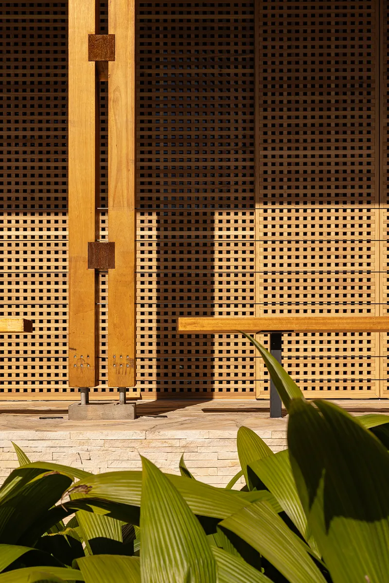 Close-up of woven lattice facade panels in shadow with timber post and tropical plant leaves below