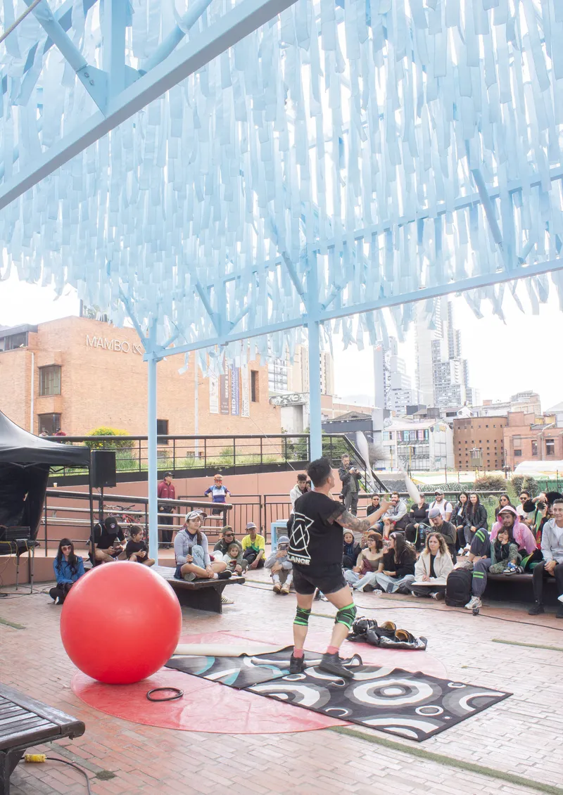 Translucent blue canopy overhead sheltering a public performance space with seated audience and large red ball