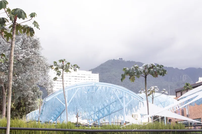 Arched canopy with translucent blue panels set among planted grasses and young trees with mountains behind