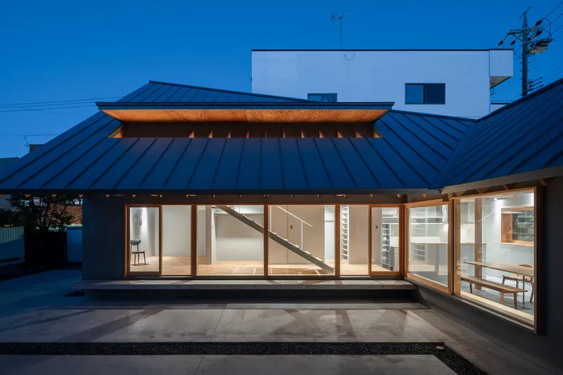 Evening view of the glazed facade beneath the metal roof with interior lighting and visible staircase