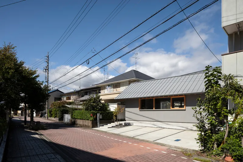 Street view showing the low-slung metal roof volumes and concrete courtyard under afternoon clouds