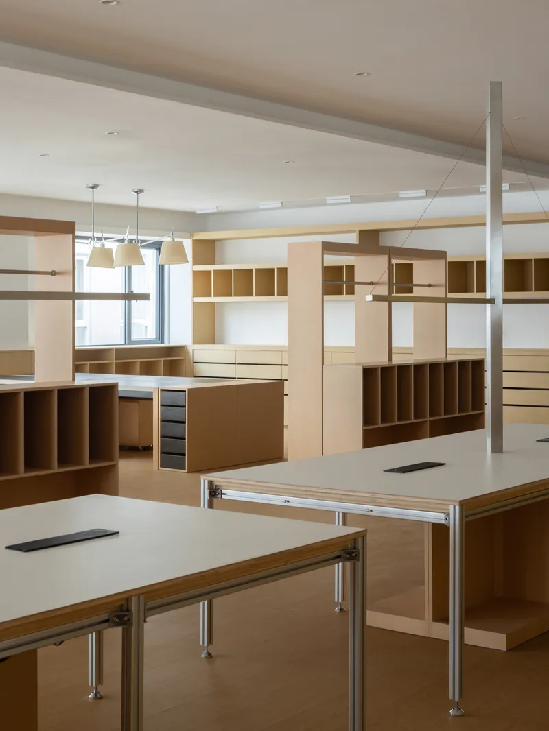 Interior workspace with plywood shelving units and white-topped tables with metal legs under pendant lights