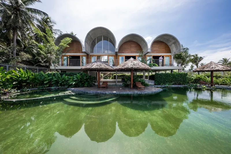Arched concrete facade with thatched pavilions reflected in a green pool surrounded by palms