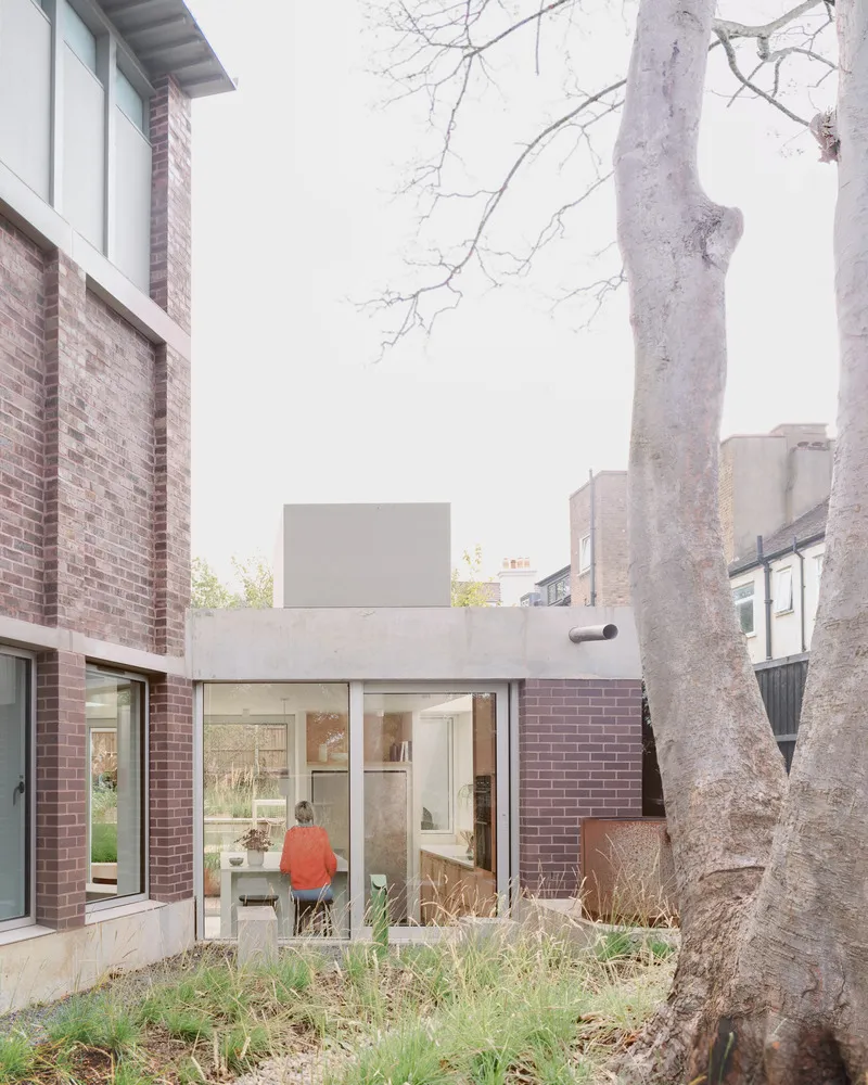 Garden view of glazed rear facade with pink brick volumes and bare tree trunk in foreground