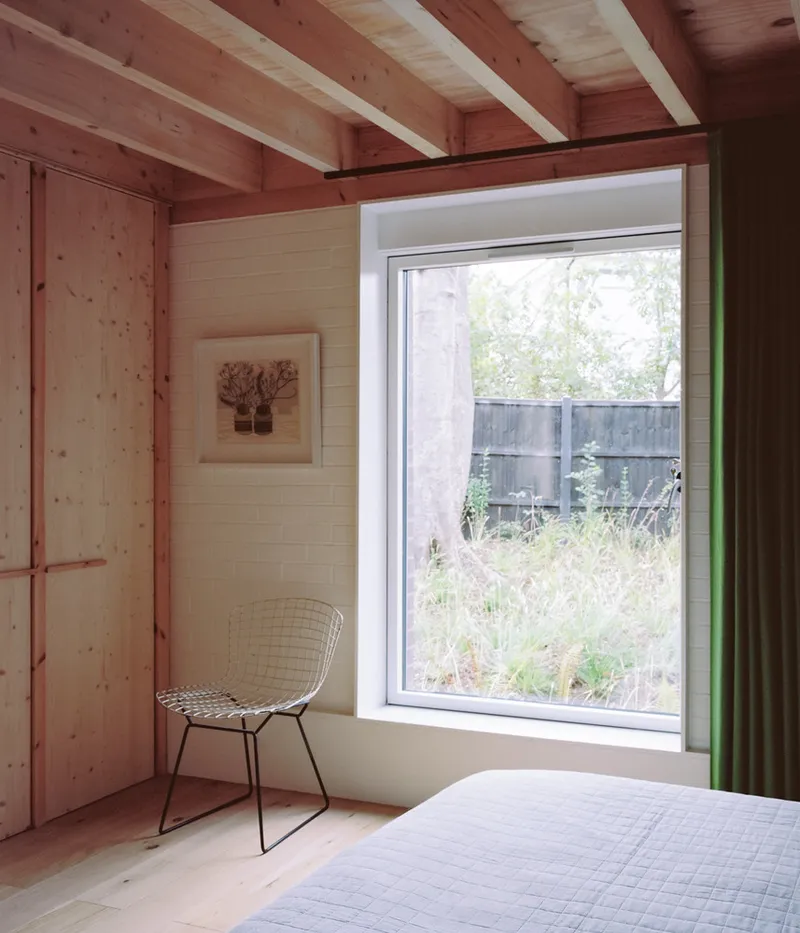 Bedroom with exposed timber beams and pale plywood walls framing a window to the garden