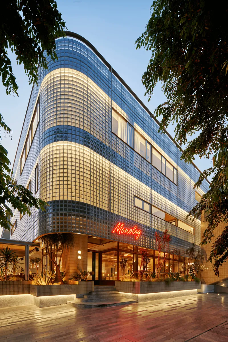Curved glass block facade rising above entrance canopy framed by tree branches at dusk