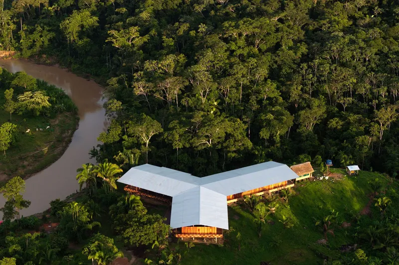 Aerial perspective of the tri-winged roof over timber volumes beside a muddy river at golden hour