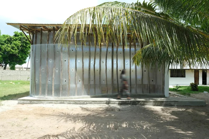 Curved corrugated metal facade with circular openings beneath overhanging palm fronds and dappled shade