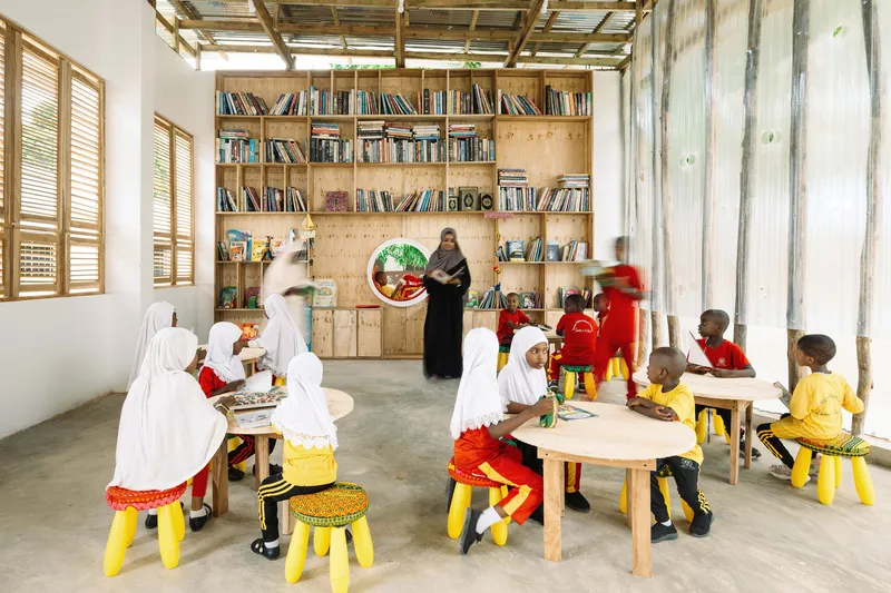 Children seated at low tables beneath exposed timber roof trusses with teacher standing by shelving wall