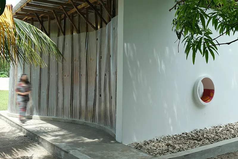 Covered walkway with vertical timber screens and a circular window opening as a figure passes through dappled light