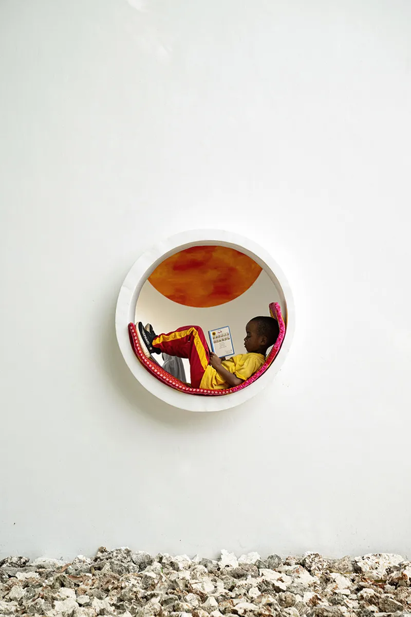 Circular opening in white wall revealing a child reading inside a nook with orange-painted interior and gravel below
