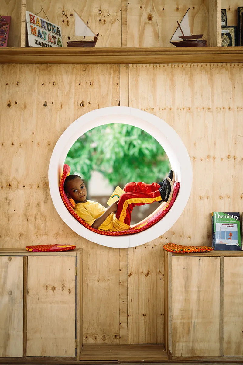 Plywood interior wall with circular reading nook containing a child looking outward toward greenery