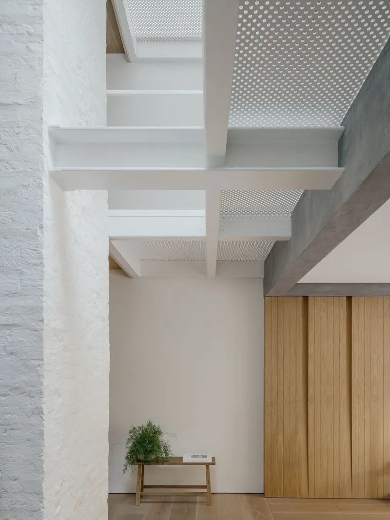 Double-height void with perforated ceiling panels, white plaster walls, and timber bench beside vertical-grain cabinetry