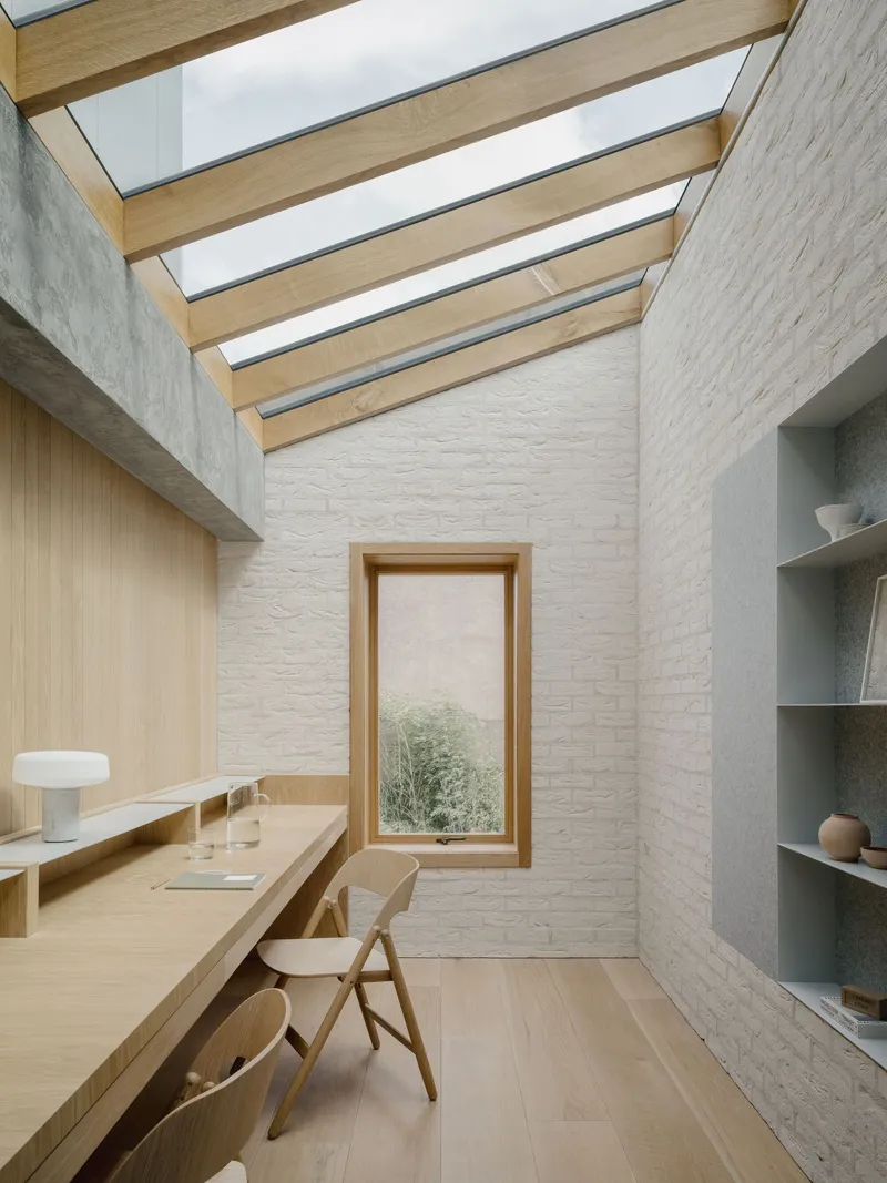 Study with timber beam skylight strips above white brick walls and vertical window overlooking greenery