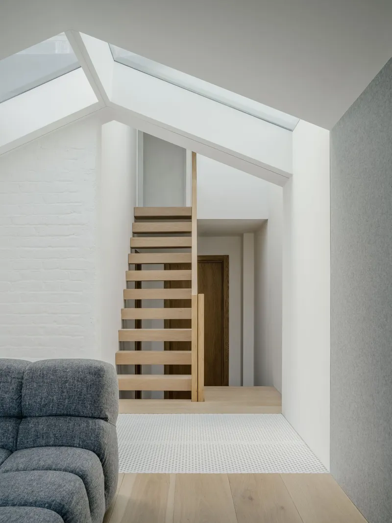Open-tread timber staircase beneath a linear skylight beside painted white brick wall